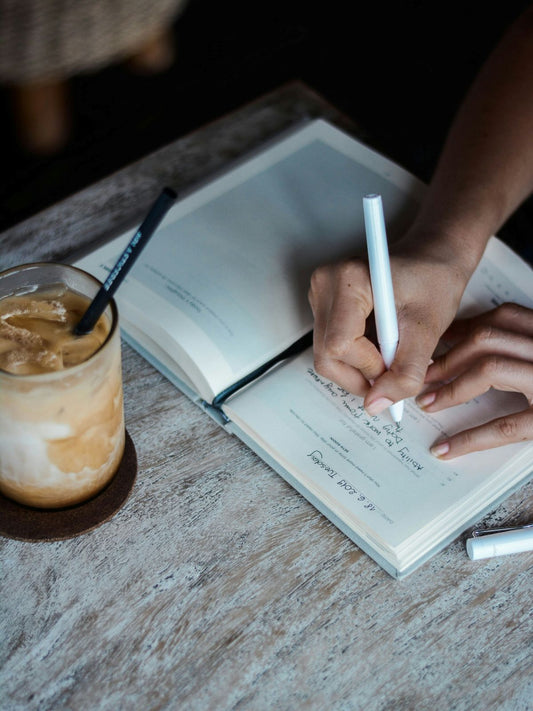 person writing in journal with iced coffee nearby